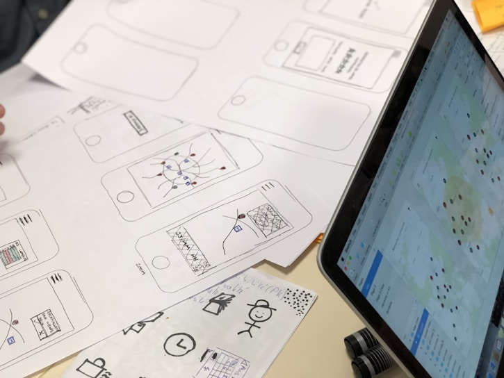A series of drawn wireframes and sketches on white paper showcasing mobile app interface designs. The designs are accompanied by a laptop displaying a digital map with various data points. Pens and other sketching tools are partially visible, indicating a workspace focused on digital design and mapping.