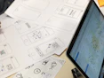 A series of drawn wireframes and sketches on white paper showcasing mobile app interface designs. The designs are accompanied by a laptop displaying a digital map with various data points. Pens and other sketching tools are partially visible, indicating a workspace focused on digital design and mapping.