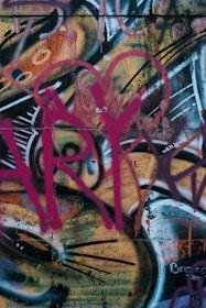 A vibrant graffiti artwork with layers of colorful spray paint, including splashes of pink, orange, black, and white. The design features abstract shapes and lines, with drips and splatters adding a dynamic feel.