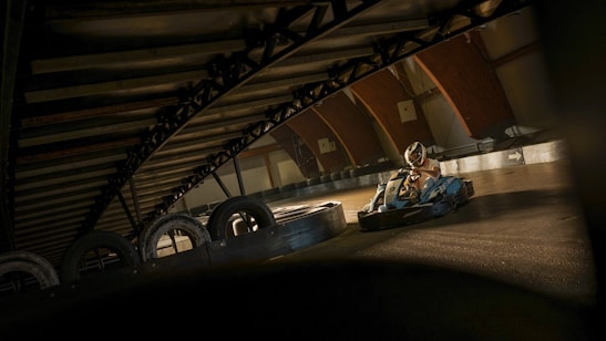A person is driving a go-kart on an indoor track with a curved, overhead metal structure. The lighting casts shadows, creating a dramatic effect. Tires are lined up as barriers on the track, and the racer is wearing protective gear.