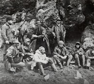 A group of men dressed in heavy coats and hats sit and stand against a rocky background. They appear to be taking a break, with some seated on the ground and others standing. Their attire suggests they are prepared for cold weather. The setting is outdoors, possibly a rugged or mountainous area.