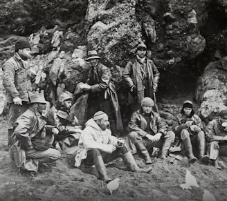 A group of men dressed in heavy coats and hats sit and stand against a rocky background. They appear to be taking a break, with some seated on the ground and others standing. Their attire suggests they are prepared for cold weather. The setting is outdoors, possibly a rugged or mountainous area.