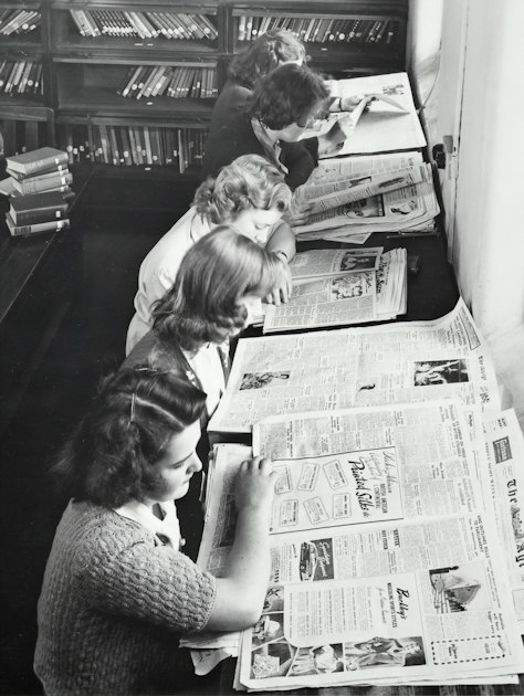 Black and white photo of three young women reading large newspapers spread out on a library reading table