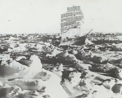 A large sailing ship navigates through a vast expanse of ice-covered waters. The scene is dominated by heavy ice floes and large masses of snow. The ship's tall masts and white sails contrast starkly with the surrounding icy landscape.