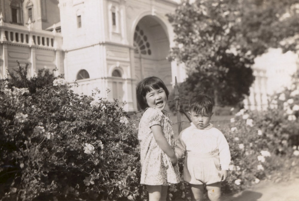 grayscale photography of girl and boy standing near flowers