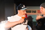 Two people are engaged in a conversation indoors. One man is holding a glass of an orange-colored cocktail and wearing a cap. The other man is looking at what might be a camera, while wearing a beanie.