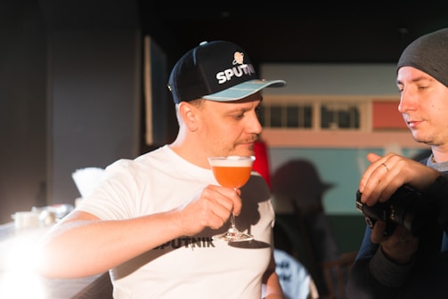 Two people are engaged in a conversation indoors. One man is holding a glass of an orange-colored cocktail and wearing a cap. The other man is looking at what might be a camera, while wearing a beanie.