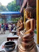 A row of golden Buddha statues is arranged on tables, with bowls placed in front of each one. People are gathered in the background, with smoke rising, possibly from incense. The setting includes a covered structure with blue and red roofing, surrounded by trees.