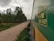 A train is traveling through a lush green landscape with trees lining the side of a dirt road. The side of the train is prominently visible, featuring text in Hindi and English about seat and sleep options. The sky is overcast with clouds, suggesting a calm, serene atmosphere.