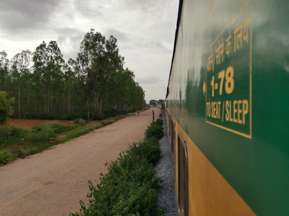 A train is traveling through a lush green landscape with trees lining the side of a dirt road. The side of the train is prominently visible, featuring text in Hindi and English about seat and sleep options. The sky is overcast with clouds, suggesting a calm, serene atmosphere.