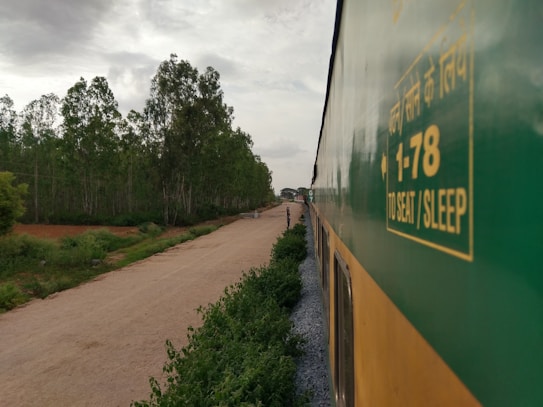 A train is traveling through a lush green landscape with trees lining the side of a dirt road. The side of the train is prominently visible, featuring text in Hindi and English about seat and sleep options. The sky is overcast with clouds, suggesting a calm, serene atmosphere.