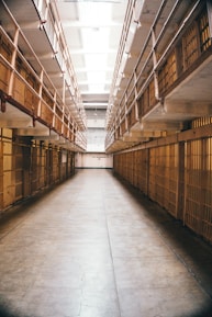 A long, narrow hallway lined with prison cells on both sides. The area is illuminated by fluorescent lights on the ceiling. Iron bars are prominently visible, creating a sense of confinement.