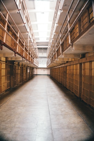 A long, narrow hallway lined with prison cells on both sides. The area is illuminated by fluorescent lights on the ceiling. Iron bars are prominently visible, creating a sense of confinement.