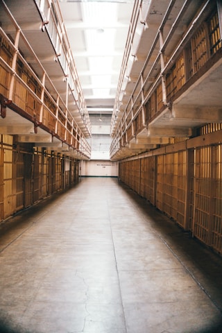 A long, narrow hallway lined with prison cells on both sides. The area is illuminated by fluorescent lights on the ceiling. Iron bars are prominently visible, creating a sense of confinement.