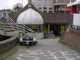 A shop with a dome-like entrance labeled 'Rijwiel Service' features bicycles displayed in front of the entrance. The setting includes an exterior staircase with metal railings and a pathway leading to the shop. In the background, there are modern buildings and several bicycles are parked nearby.