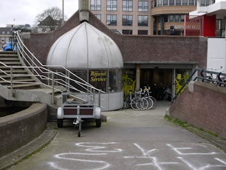 A shop with a dome-like entrance labeled 'Rijwiel Service' features bicycles displayed in front of the entrance. The setting includes an exterior staircase with metal railings and a pathway leading to the shop. In the background, there are modern buildings and several bicycles are parked nearby.