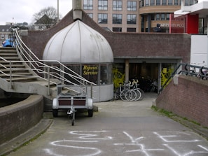 A shop with a dome-like entrance labeled 'Rijwiel Service' features bicycles displayed in front of the entrance. The setting includes an exterior staircase with metal railings and a pathway leading to the shop. In the background, there are modern buildings and several bicycles are parked nearby.