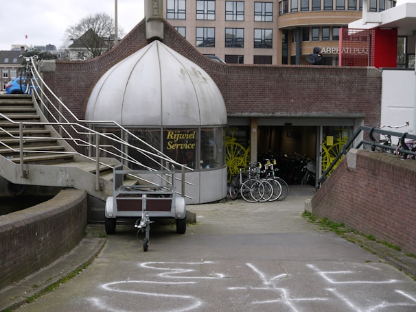 A shop with a dome-like entrance labeled 'Rijwiel Service' features bicycles displayed in front of the entrance. The setting includes an exterior staircase with metal railings and a pathway leading to the shop. In the background, there are modern buildings and several bicycles are parked nearby.