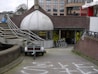 A shop with a dome-like entrance labeled 'Rijwiel Service' features bicycles displayed in front of the entrance. The setting includes an exterior staircase with metal railings and a pathway leading to the shop. In the background, there are modern buildings and several bicycles are parked nearby.