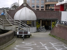 A shop with a dome-like entrance labeled 'Rijwiel Service' features bicycles displayed in front of the entrance. The setting includes an exterior staircase with metal railings and a pathway leading to the shop. In the background, there are modern buildings and several bicycles are parked nearby.