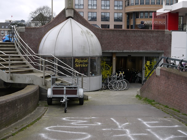 A shop with a dome-like entrance labeled 'Rijwiel Service' features bicycles displayed in front of the entrance. The setting includes an exterior staircase with metal railings and a pathway leading to the shop. In the background, there are modern buildings and several bicycles are parked nearby.