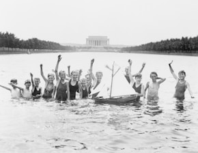 A group of children are joyfully playing in a shallow pool of water with a model sailboat floating nearby. In the background, the Lincoln Memorial is visible, and the area is lined with trees. The children are waving and smiling, creating a lively, cheerful atmosphere.