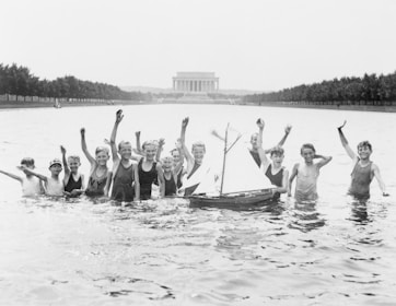 A group of children are joyfully playing in a shallow pool of water with a model sailboat floating nearby. In the background, the Lincoln Memorial is visible, and the area is lined with trees. The children are waving and smiling, creating a lively, cheerful atmosphere.