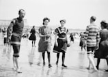 A group of people dressed in early 20th-century beach attire are walking along a sandy shore. The image captures men and women wearing vintage swimsuits with high collars and stripes, typical of the era. The beach is crowded, suggesting a popular coastal location during a warm day.
