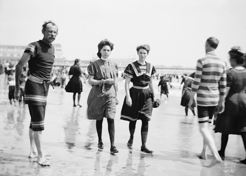 A group of people dressed in early 20th-century beach attire are walking along a sandy shore. The image captures men and women wearing vintage swimsuits with high collars and stripes, typical of the era. The beach is crowded, suggesting a popular coastal location during a warm day.