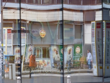 The image captures a reflection in a glass window of an urban street scene. People are walking past storefronts with colorful advertisements. The word 'LOVE' is visible in green outlined letters on the glass, along with other words that are partially obscured. Buildings and signs are distorted by the glass reflections.