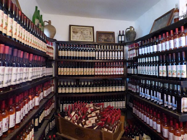 A room filled with shelves stacked with a variety of wine bottles. The bottles have different labels and are neatly organized. The shelves are arranged along the walls, creating an impression of abundance. In the center, an open wooden crate holds additional bottles. The room has framed pictures and some decorative clay and glass containers on top of the shelves.