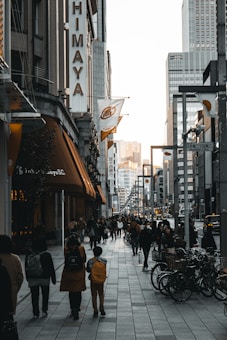 A busy urban street scene featuring numerous pedestrians walking along the sidewalk. The street is lined with tall buildings, and there are bicycles parked to the side. The lighting suggests it is either early morning or late afternoon, adding a warm hue to the scene. Signage with vertical and horizontal text in an Asian script is visible.