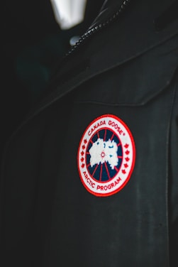 A close-up of a garment featuring a circular patch with a red border, showcasing a stylized map of the Arctic. The patch reads 'Canada Goose Arctic Program' and is sewn onto a dark fabric, possibly a jacket or coat.