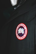 A close-up of a garment featuring a circular patch with a red border, showcasing a stylized map of the Arctic. The patch reads 'Canada Goose Arctic Program' and is sewn onto a dark fabric, possibly a jacket or coat.
