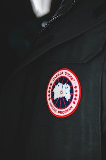 A close-up of a garment featuring a circular patch with a red border, showcasing a stylized map of the Arctic. The patch reads 'Canada Goose Arctic Program' and is sewn onto a dark fabric, possibly a jacket or coat.