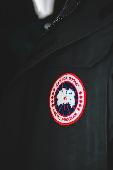 A close-up of a garment featuring a circular patch with a red border, showcasing a stylized map of the Arctic. The patch reads 'Canada Goose Arctic Program' and is sewn onto a dark fabric, possibly a jacket or coat.