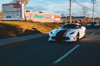 A sleek white sports car with black and red racing stripes is driving down a road. The sun casts long shadows, and there are a few other vehicles on the road. A brick building with the Sobeys logo is visible along with a pharmacy sign. The surrounding area includes trees and mountains in the background, and power lines run along the road.