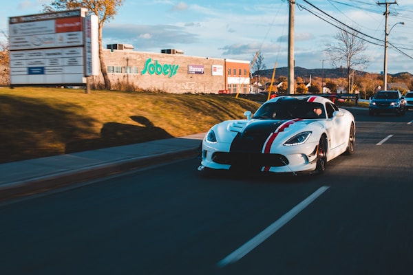 A sleek white sports car with black and red racing stripes is driving down a road. The sun casts long shadows, and there are a few other vehicles on the road. A brick building with the Sobeys logo is visible along with a pharmacy sign. The surrounding area includes trees and mountains in the background, and power lines run along the road.