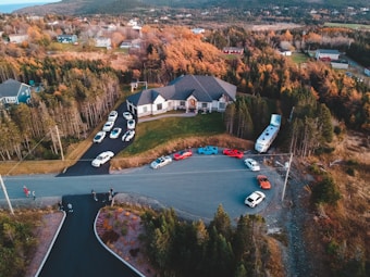 A large house is surrounded by a group of sports cars parked on a circular driveway. The property is nestled among trees with autumn foliage. Several people are standing near the cars and on the road leading up to the house. A large RV is parked near the house, and more residential buildings and dense forest cover the distant hills.