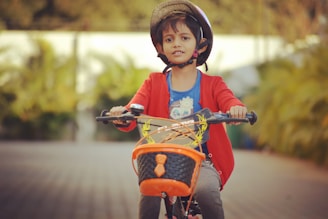 A young child wearing a helmet and a red sweater is riding a bicycle with an orange basket. The background is a blurred pathway with greenery on the sides.