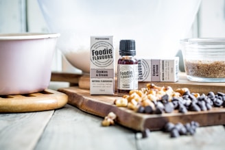 A bottle and packaging of cookies and cream flavoring are placed on a wooden cutting board surrounded by various baking ingredients, including chocolate chips and nuts. A mixing bowl is visible in the background, along with a pink mixing bowl and a glass dish filled with sugar or a similar ingredient.