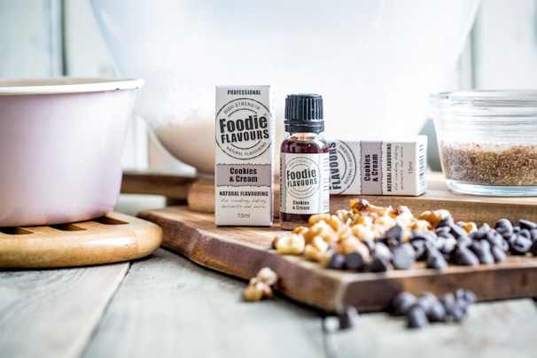 A bottle and packaging of cookies and cream flavoring are placed on a wooden cutting board surrounded by various baking ingredients, including chocolate chips and nuts. A mixing bowl is visible in the background, along with a pink mixing bowl and a glass dish filled with sugar or a similar ingredient.