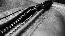 A close-up view of a leather surface with a prominent metal zipper. The texture of the leather is detailed and slightly shiny, and the zipper is partially open, with the teeth interlocking.