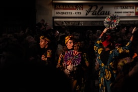A group of people dressed in vibrant, traditional clothing stand in an area that suggests a festive or performance setting. They hold decorative fans and have elaborate headwear, with the lighting highlighting their colorful outfits against the dark background. A sign in the background reads 'Scalextric Palau Maquetes - Kits'.