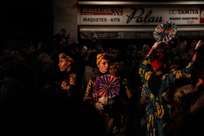 A group of people dressed in vibrant, traditional clothing stand in an area that suggests a festive or performance setting. They hold decorative fans and have elaborate headwear, with the lighting highlighting their colorful outfits against the dark background. A sign in the background reads 'Scalextric Palau Maquetes - Kits'.