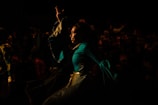 A person dressed in a vibrant, flowing costume performing on stage or in a theatrical setting. The background is dark, with a crowd of people faintly visible, suggesting a lively performance or festival atmosphere.