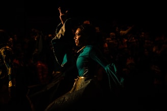 A person dressed in a vibrant, flowing costume performing on stage or in a theatrical setting. The background is dark, with a crowd of people faintly visible, suggesting a lively performance or festival atmosphere.