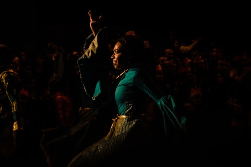 A person dressed in a vibrant, flowing costume performing on stage or in a theatrical setting. The background is dark, with a crowd of people faintly visible, suggesting a lively performance or festival atmosphere.