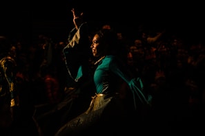 A person dressed in a vibrant, flowing costume performing on stage or in a theatrical setting. The background is dark, with a crowd of people faintly visible, suggesting a lively performance or festival atmosphere.