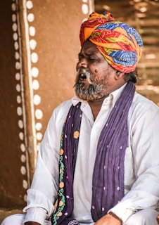 A man wearing a colorful turban and traditional attire is captured mid-expression, possibly singing or speaking passionately. The background features rustic, earthy tones and circular patterns.